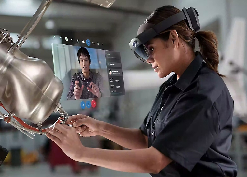 Một người phụ nữ đeo mắt kính thực tế ảo làm việc trên bộ phận cơ khí trong nhà máy, tương tác với màn hình ảo.