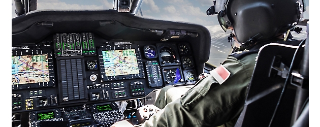 Pilot in military helicopter cockpit with detailed instrument panels and multiple display screens.