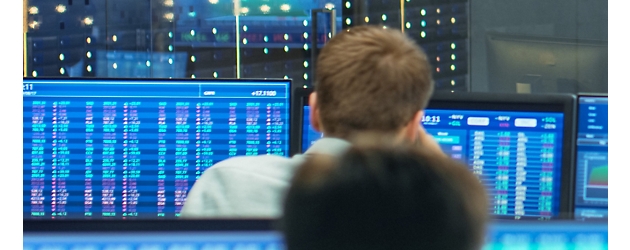 Vista traseira de duas pessoas a monitorizar dados financeiros em vários ecrãs azuis num ambiente de sala de controlo.