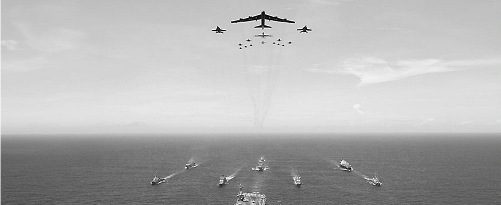 Aerial view of a fleet of ships on the ocean with a formation of military aircraft flying above, leaving trails in the sky.