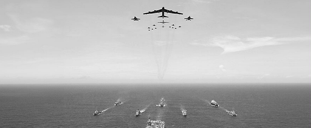Aerial view of a fleet of ships on the ocean with a formation of military aircraft flying above, leaving trails in the sky.