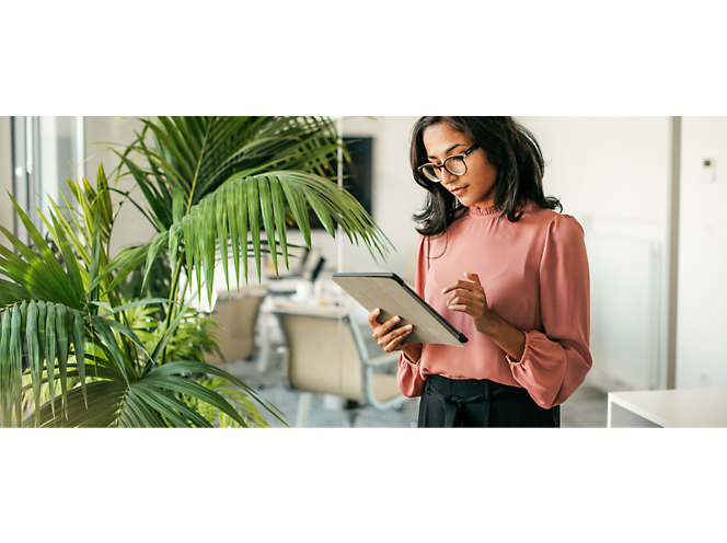 Una mujer profesional con blusa rosa y gafas leyendo en una tableta en una oficina moderna con una gran planta en una maceta.