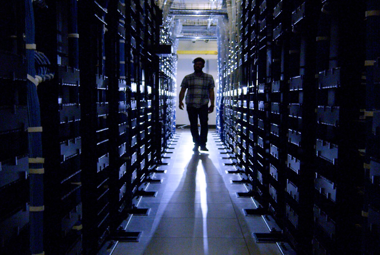 A man walks through a dimly lit hallway between rows of server racks in a data center.