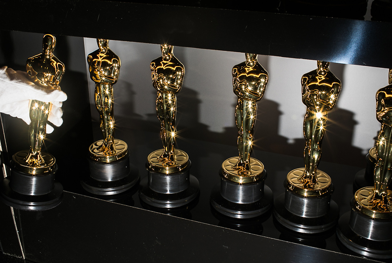 A gloved hand places an Oscar statuette on a black shelf among five other identical statues.