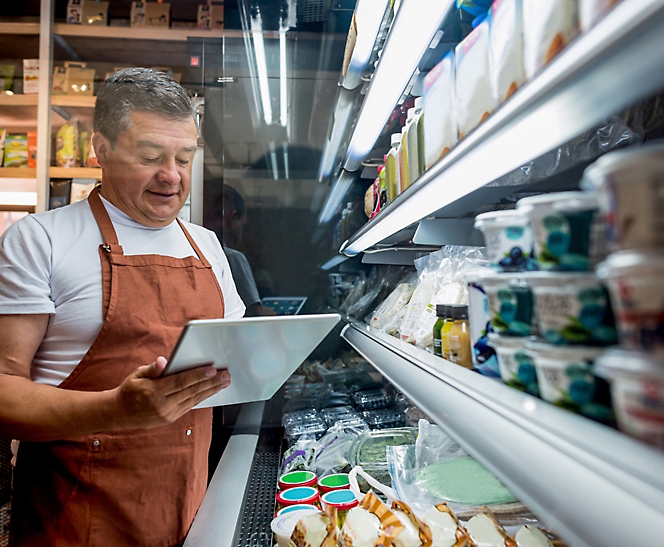 A store employee in an apron checks inventory with a tablet in front of a dairy refrigerator.