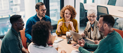 A diverse group of six professionals engaged in a meeting around a table, with one person showing a tablet to others.