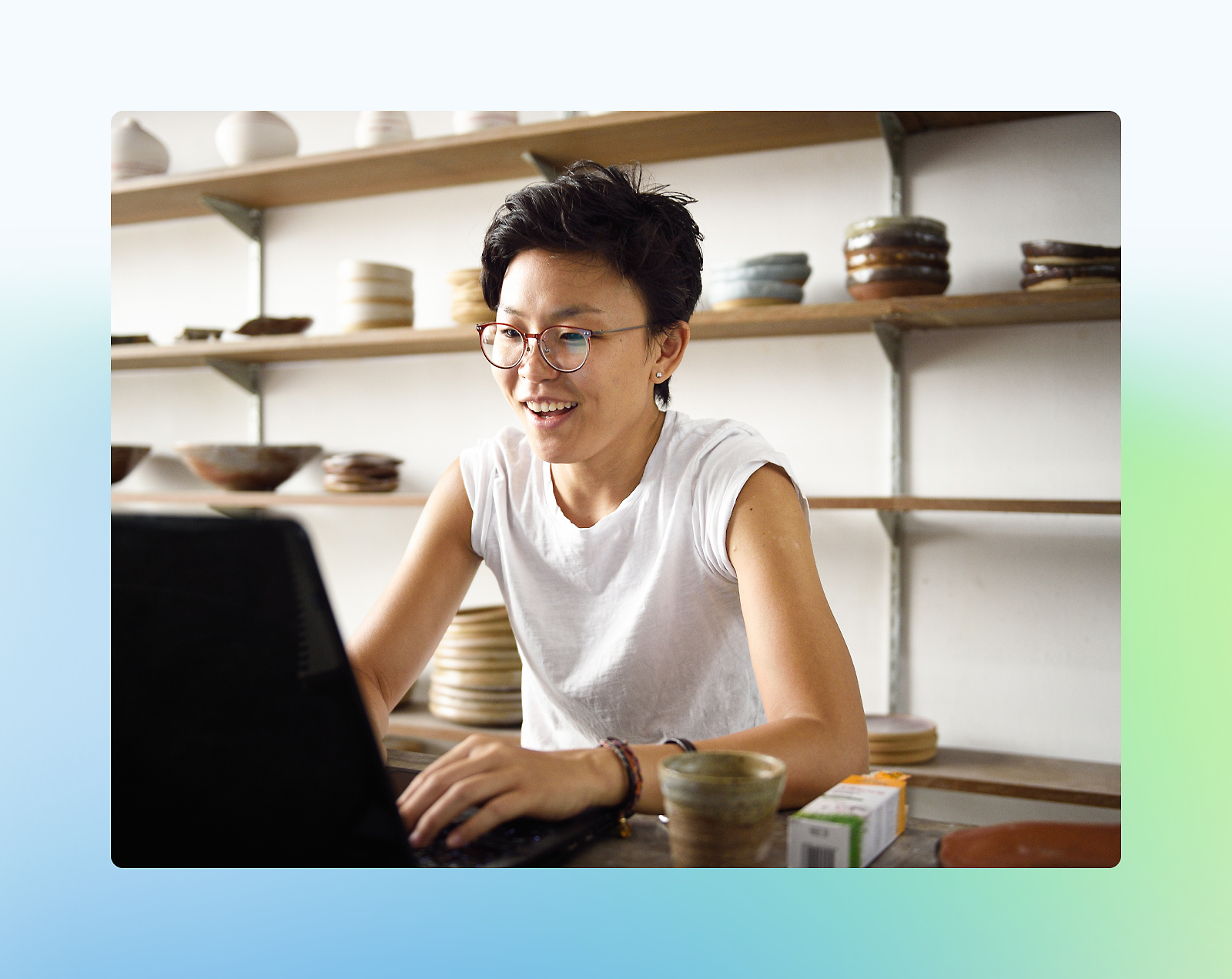 Eine Person, die eine Brille und ein weißes T-Shirt trägt, lächelt, während sie einen Laptop in einem Raum mit Regalen voller Keramik verwendet.