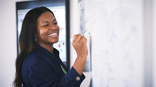 A smiling woman writing on a whiteboard in an office setting.