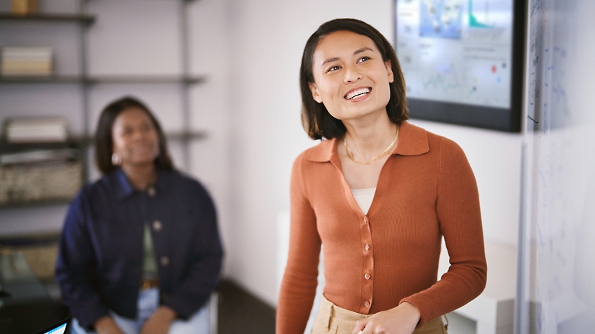 Due donne in ufficio, una in piedi in primo piano sorridente verso la fotocamera e un'altra seduta sullo sfondo.