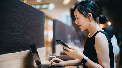 A woman in a black sleeveless top is sitting at a desk, holding a smartphone in her right hand and typing on a laptop.