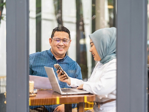 Twee mensen zitten in een café, lachend en werkend aan een project. De ene persoon in een blauwe blouse bevat een document, terwijl de andere persoon een hijab draagt.