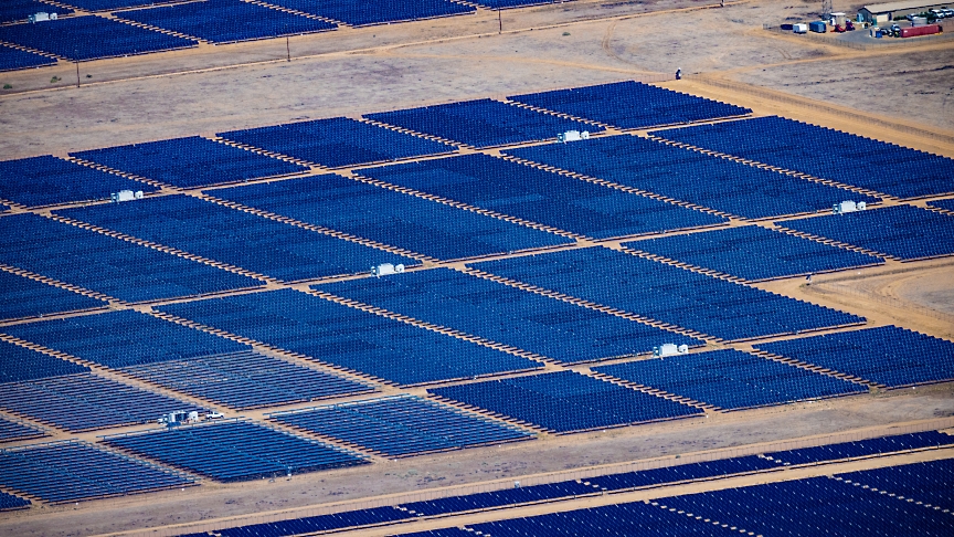 Vista aérea de um grande parque solar com filas de painéis solares azuis ordenadamente dispostos numa paisagem árida.