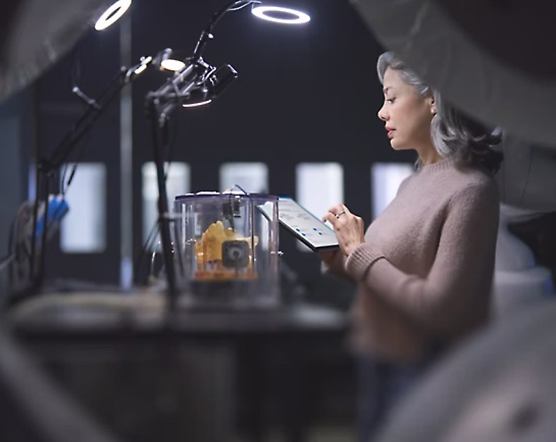 Eine Frau prüft einen 3D-Druck, während sie ein Tablet in einer Workshop-Umgebung hält.
