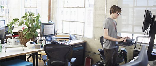 Osoba w okularach pracująca w biurze, która stoi używając komputera stacjonarnego