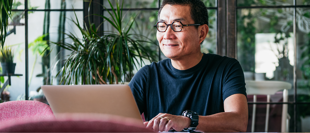 Hombre asiático de mediana edad sonriendo y apartando la mirada de su portátil en una cafetería.