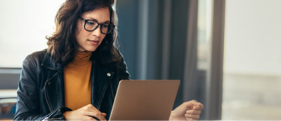 Woman in glasses and a leather jacket working intently on a laptop near a window.