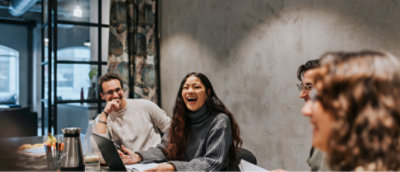Three people enjoying a conversation at a table in a cafe, one woman laughing joyfully with a laptop open in front of them.