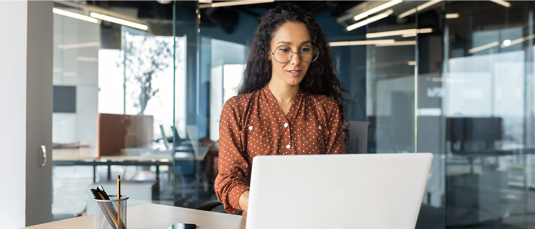 Kobieta w okularach z kręconymi włosami, ubrana w bluzkę w kropki typu polka, która pracuje na laptopie w nowoczesnym biurze.