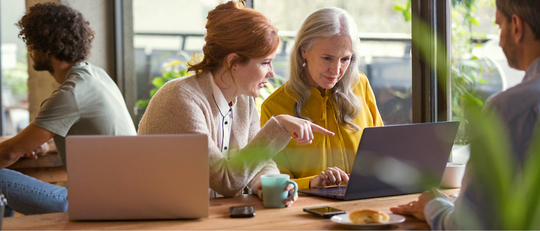 Duas mulheres, uma mais jovem e outra mais velha, estão olhando juntas para a tela de um laptop em um café durante o dia.