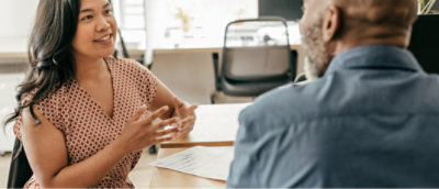 A woman and a man conversing over a document at a table in a brightly lit office setting.