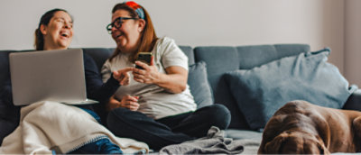 Two women laughing and sitting on a couch with a laptop and a smartphone, a dog lying nearby.