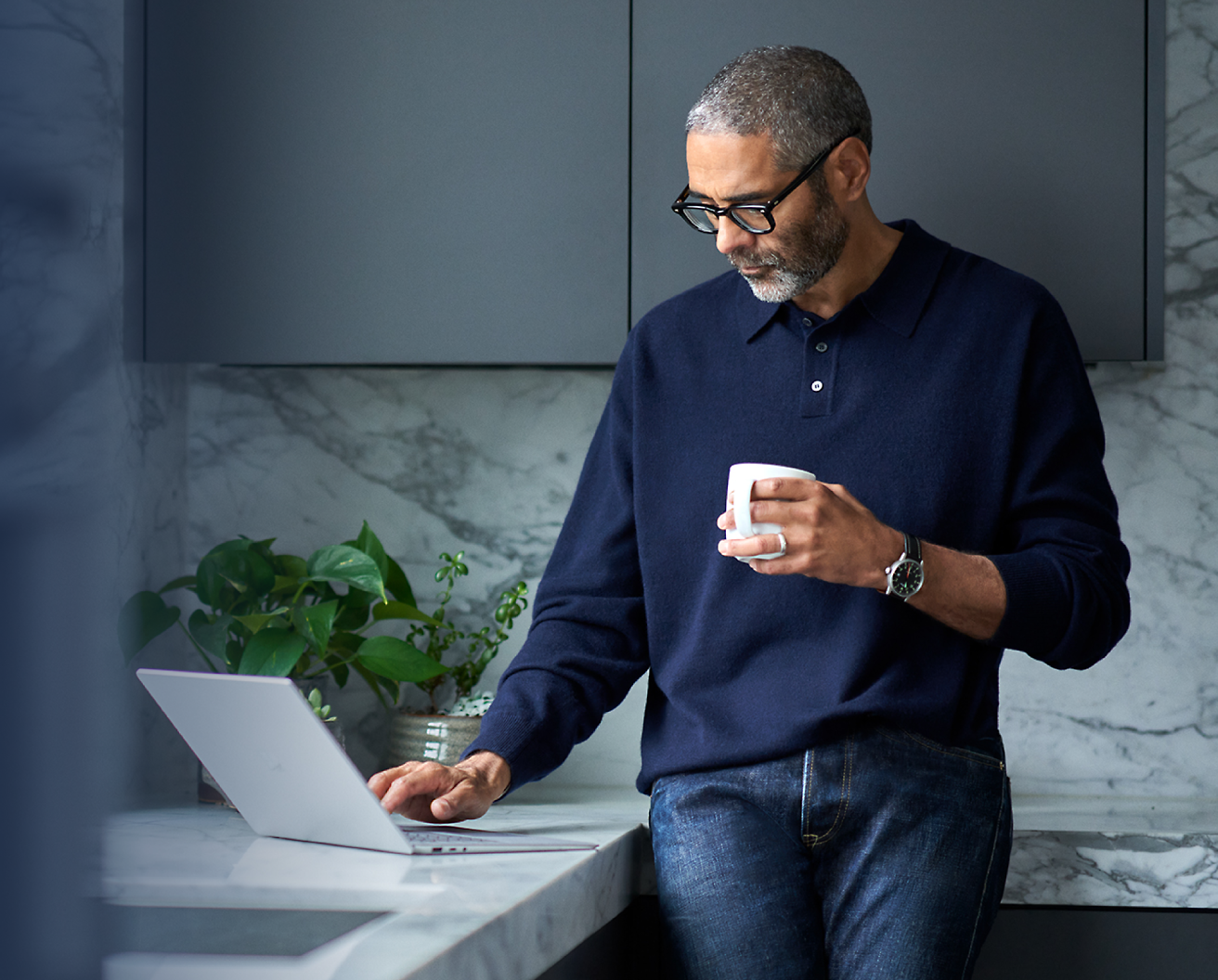 Ein Mann mittleren Alters mit Brille in einer modernen Küche, der eine Kaffeetasse hält und einen Laptop verwendet