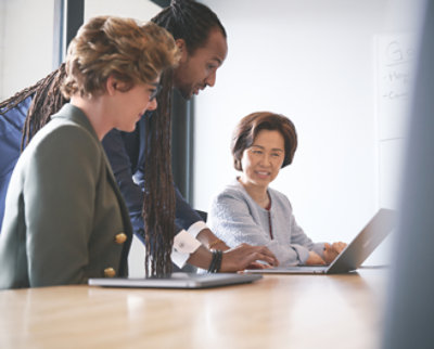 Three professionals discussing over a laptop at a conference table in a bright office, with a whiteboard visible in the background.