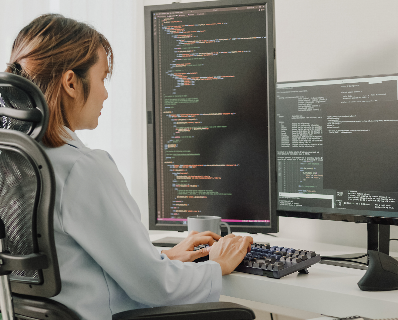Uma mulher sentada em uma cadeira de escritório, programando em um computador com várias telas exibindo código de programação.