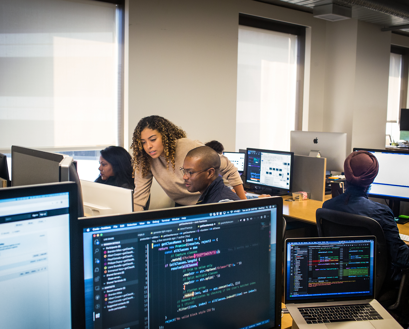 En mangfoldig gruppe fagpersoner arbejder på computere i et kontormiljø, hvor de fokuserer på skærme med forskellig synlig kode og data.