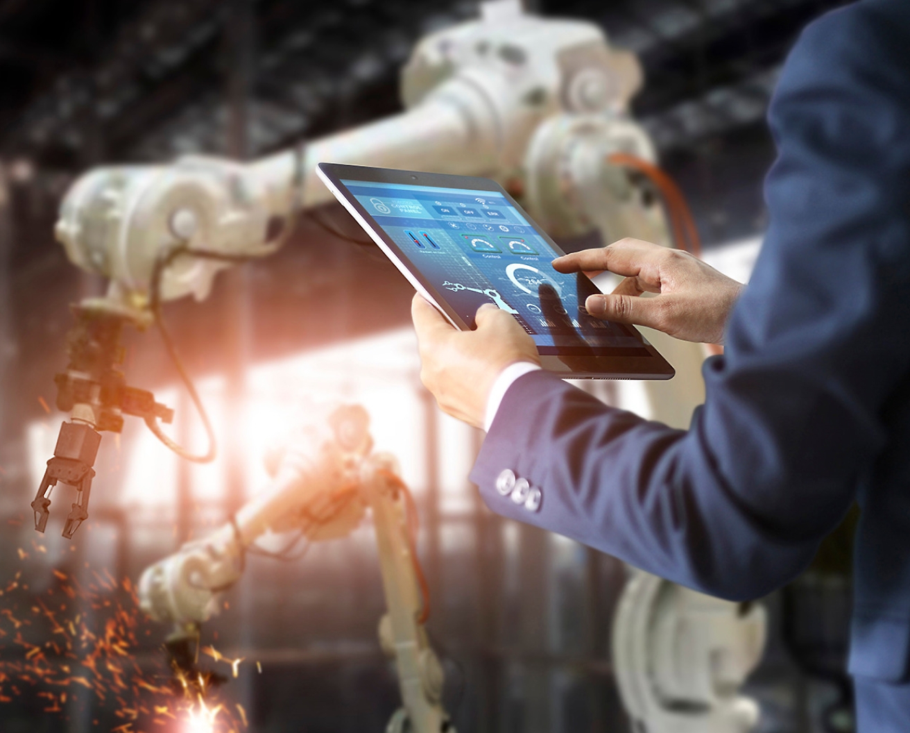 Osoba v továrně ovládá robotická ramena pomocí tabletu, což poukazuje na pokročilou automatizační technologii