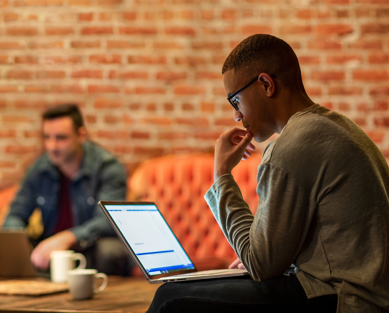 Um homem focado, usando óculos, trabalha em um laptop em um ambiente de escritório casual, com outra pessoa ao fundo.