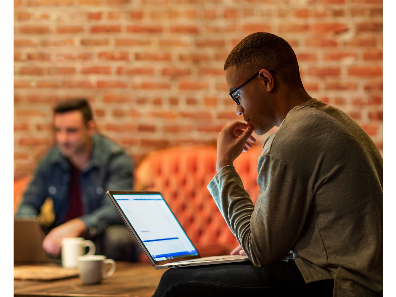 Ein konzentrierter Mann mit Brille benutzt einen Laptop in einer lockeren Büroumgebung, während im Hintergrund eine weitere Person zu sehen ist.