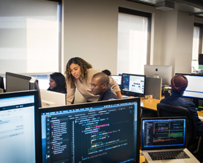 Diverse group of professionals working on computers in an office environment, focusing on screens with various code and data visible.