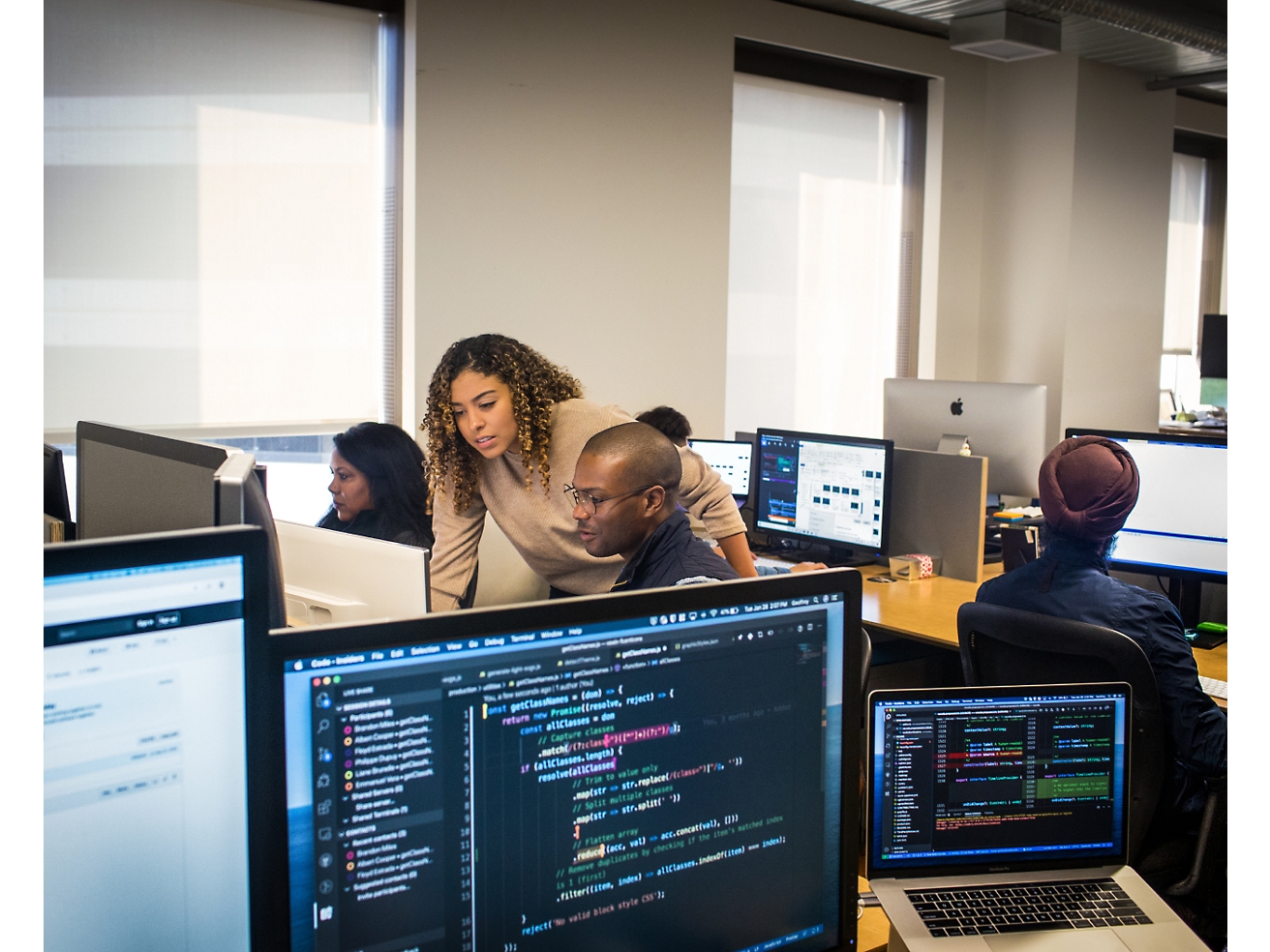 Группа разнообразных специалистов работает за компьютерами в офисной обстановке, глядя на экраны, на которых отображается различный программный код и данные.