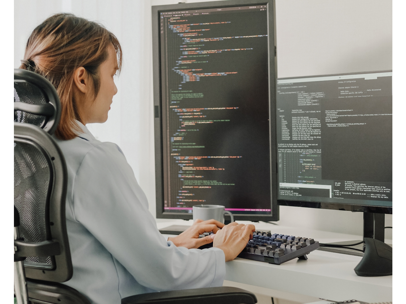 オフィス チェアに座り、プログラミング コードを表示する複数のスクリーン ディスプレイのあるコンピューターでコーディングをしている女性。