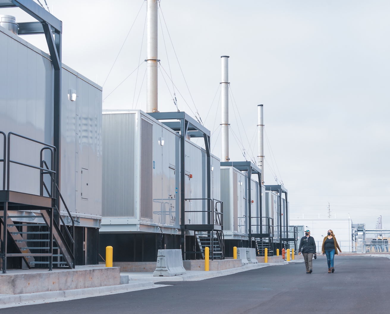 Two people walking by a row of industrial containers with tall chimneys at a facility, under an overcast sky.