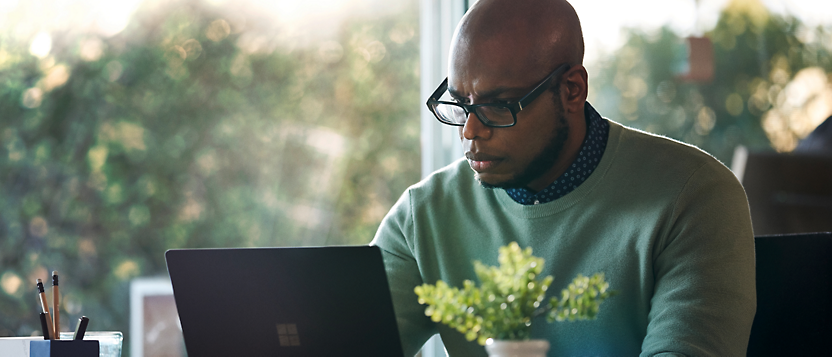 Un uomo concentrato con occhiali lavora su un portatile alla scrivania, con uno sfondo con luce soffusa e una piccola pianta vicino a lui.