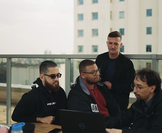 四位男士（其中一人使用笔记本电脑）在一张户外桌子上进行严肃的讨论，背景是城市建筑。