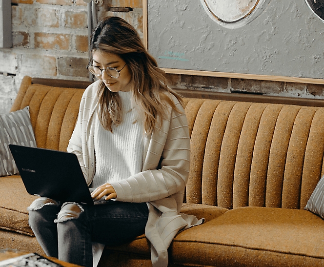 A woman in glasses and a striped sweater sits on a mustard couch, working on a laptop in a cozy café setting.