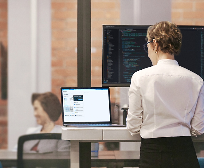 Kobieta stoi przy wysokim biurku i pracuje na laptopie z dużym monitorem wyświetlającym kod w tle.