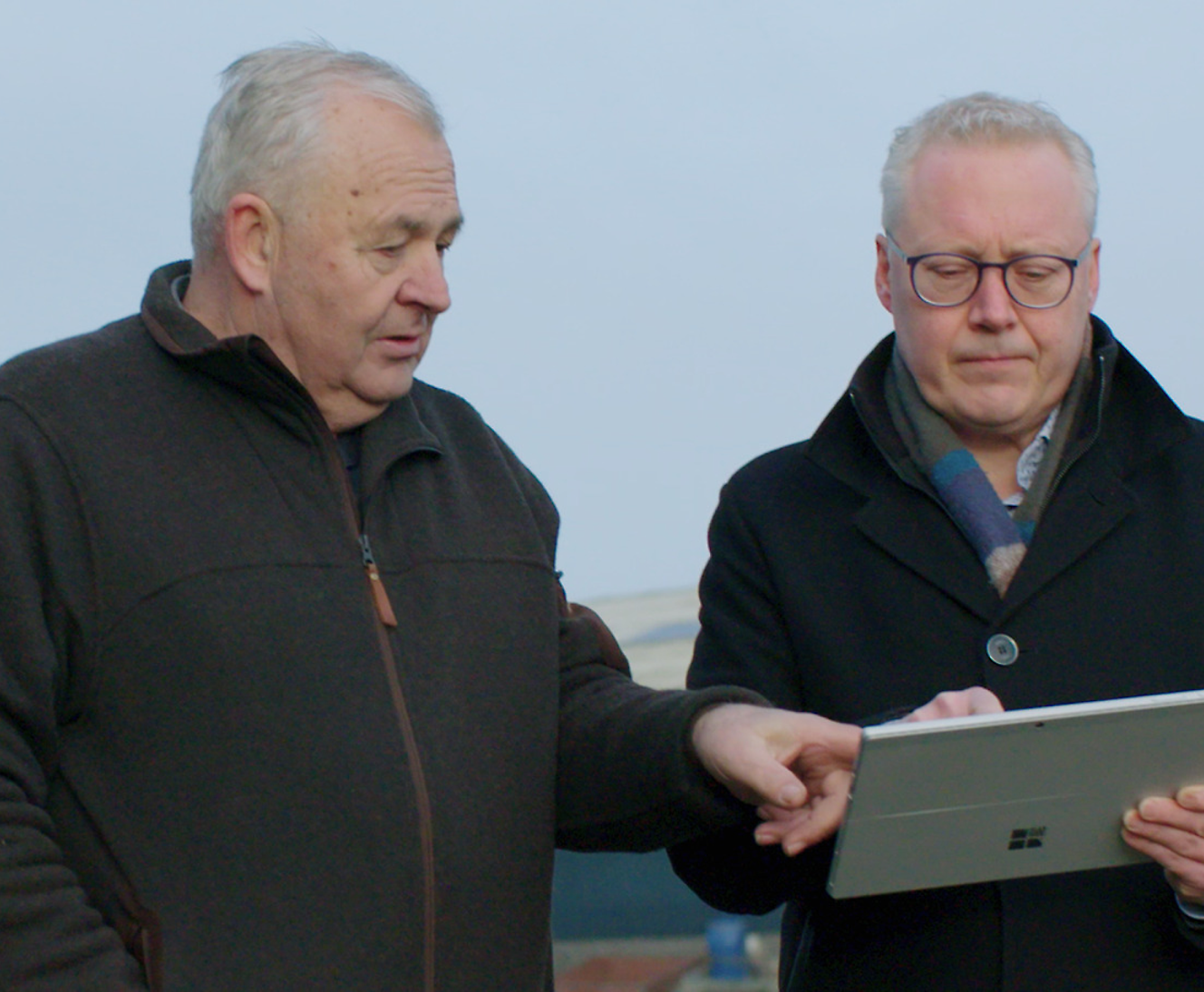 Two individuals stand outdoors, with one man using a tablet while the other points at the screen.