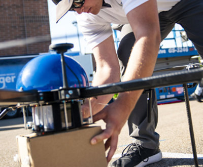 En person i en vit skjorta och gråa byxor arbetar på en stor drönare och skyddar ett paket under den