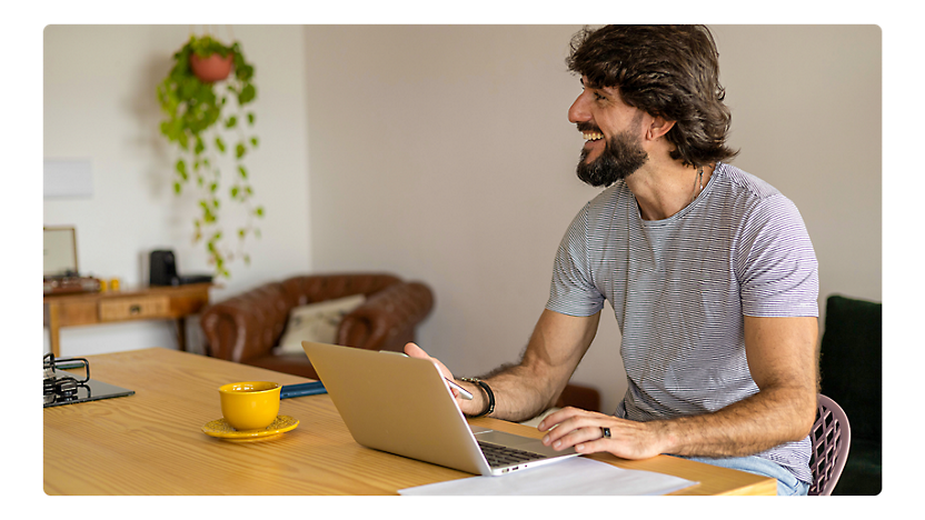A man with a beard smiling while using a laptop at a home office desk with a cup of coffee and plant decor.