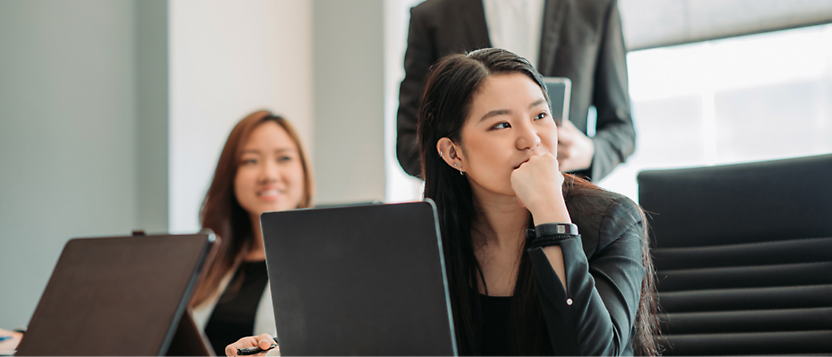 A young Asian woman in business attire listens intently in a meeting, with a laptop open and another attendee 