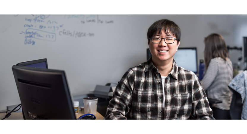 En smilende østasiatisk person med briller og ternet skjorte sidder foran en computer på et kontor med et whiteboard 