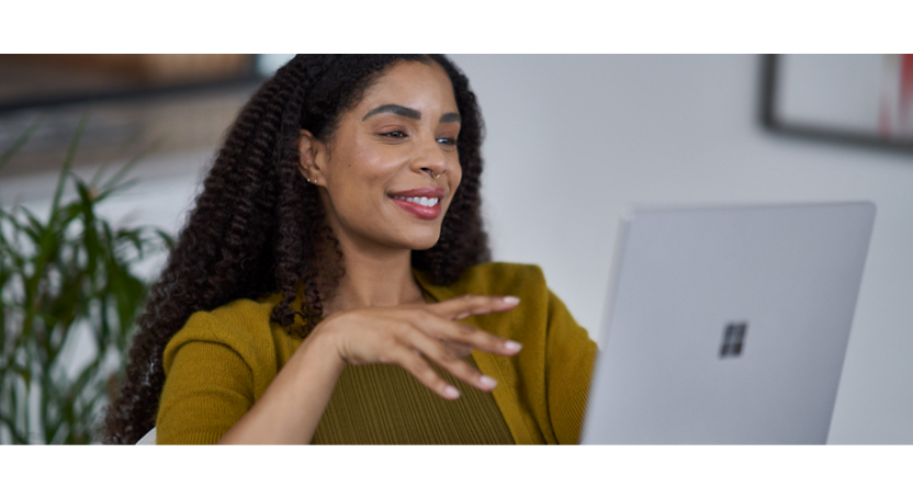 Egy göndör hajú nő finoman mosolyog, miközben a laptop képernyőjét nézi, mustársárga pulóvert visel.