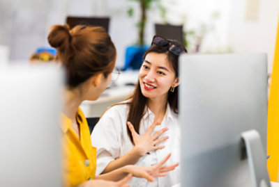Twee vrouwen in gesprek op het werk, de een in een geel shirt en de ander in een wit shirt met een bril op haar hoofd.
