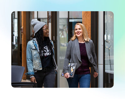 2 人の女性が屋内で歩きながら話している。1 人はビーニー帽とデニム ジャケットを身に付けており、もう 1 人はノート PC を持っているブロンド ヘアの女性。