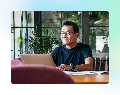 デスクに座ってノート PC で仕事をしている、眼鏡をかけて黒いシャツを着ている男性。