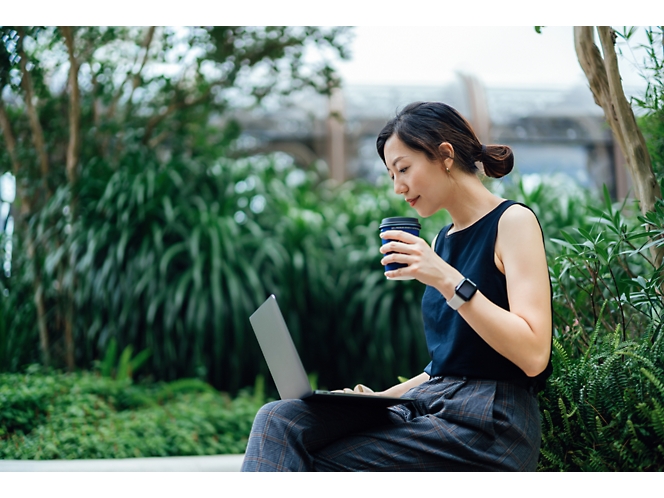 A woman sits outdoors on a bench with foliage behind her, holding a coffee cup in one hand and using a laptop